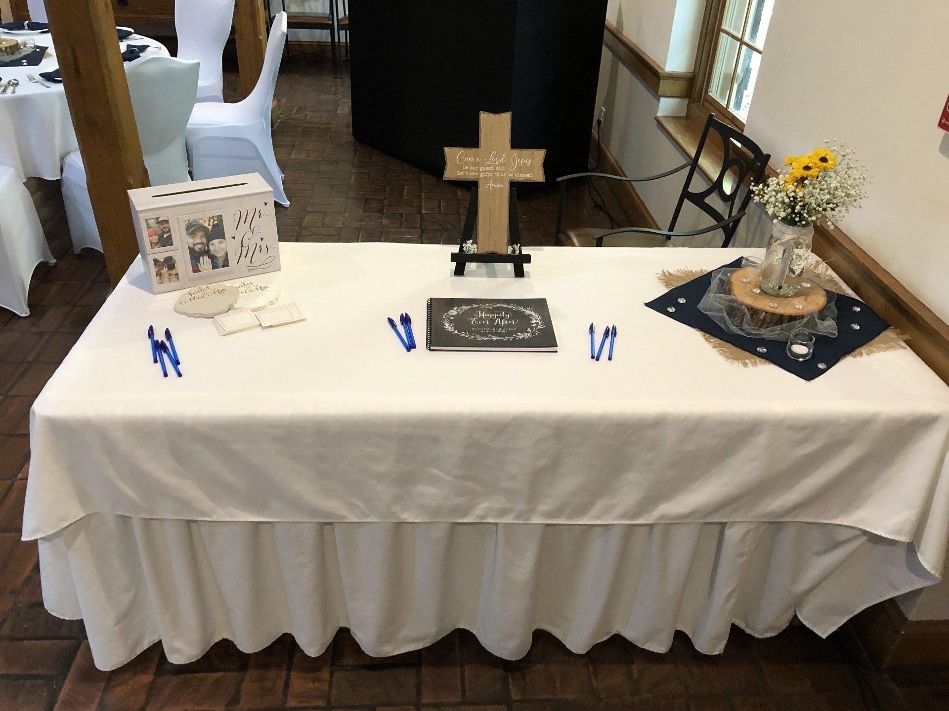 A guest book table with a white tablecloth, cross, donation box, pens, and decorations in a room.