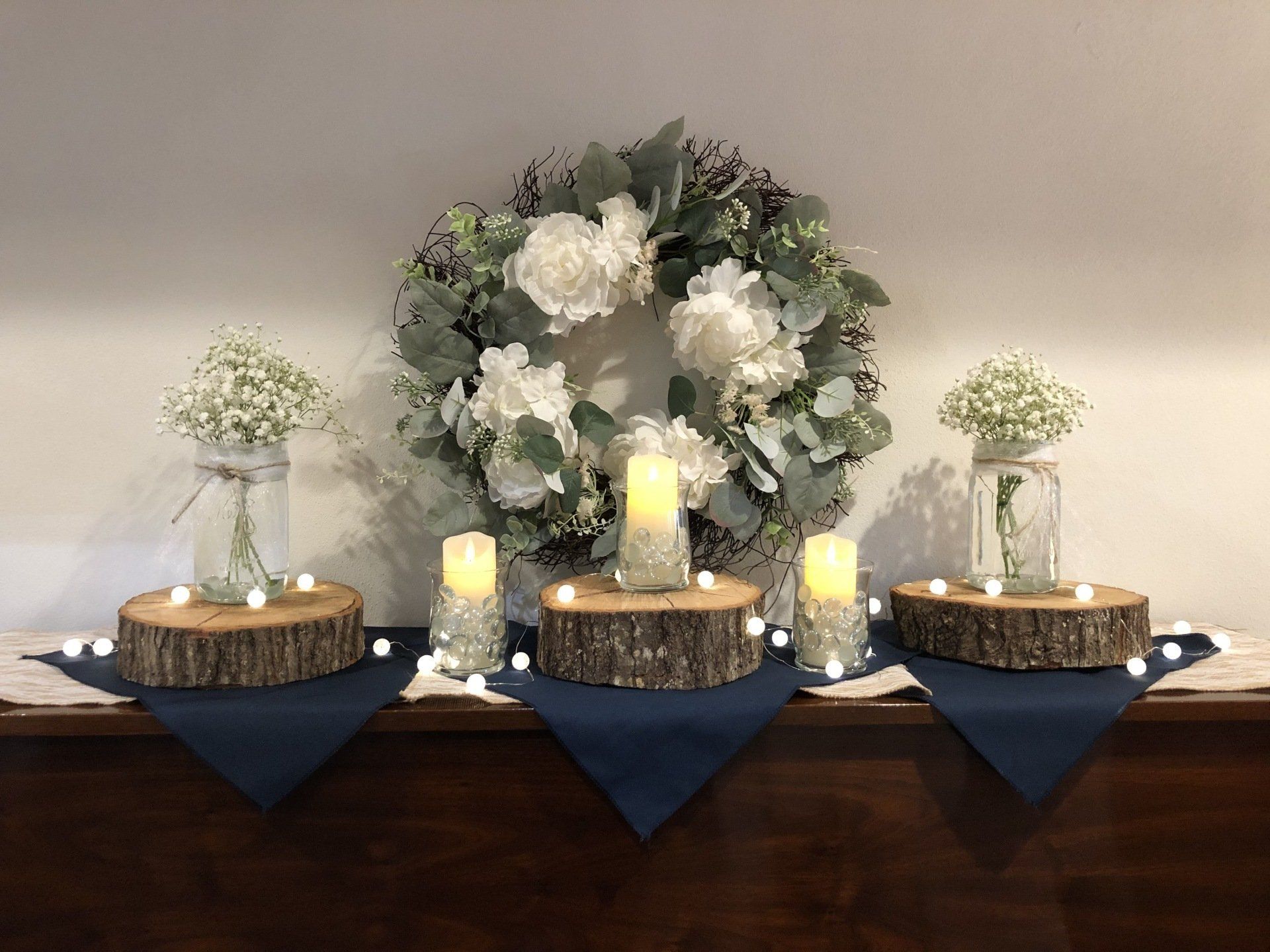 Display with white flowers, candles, wood slices, and a wreath on a navy blue cloth.