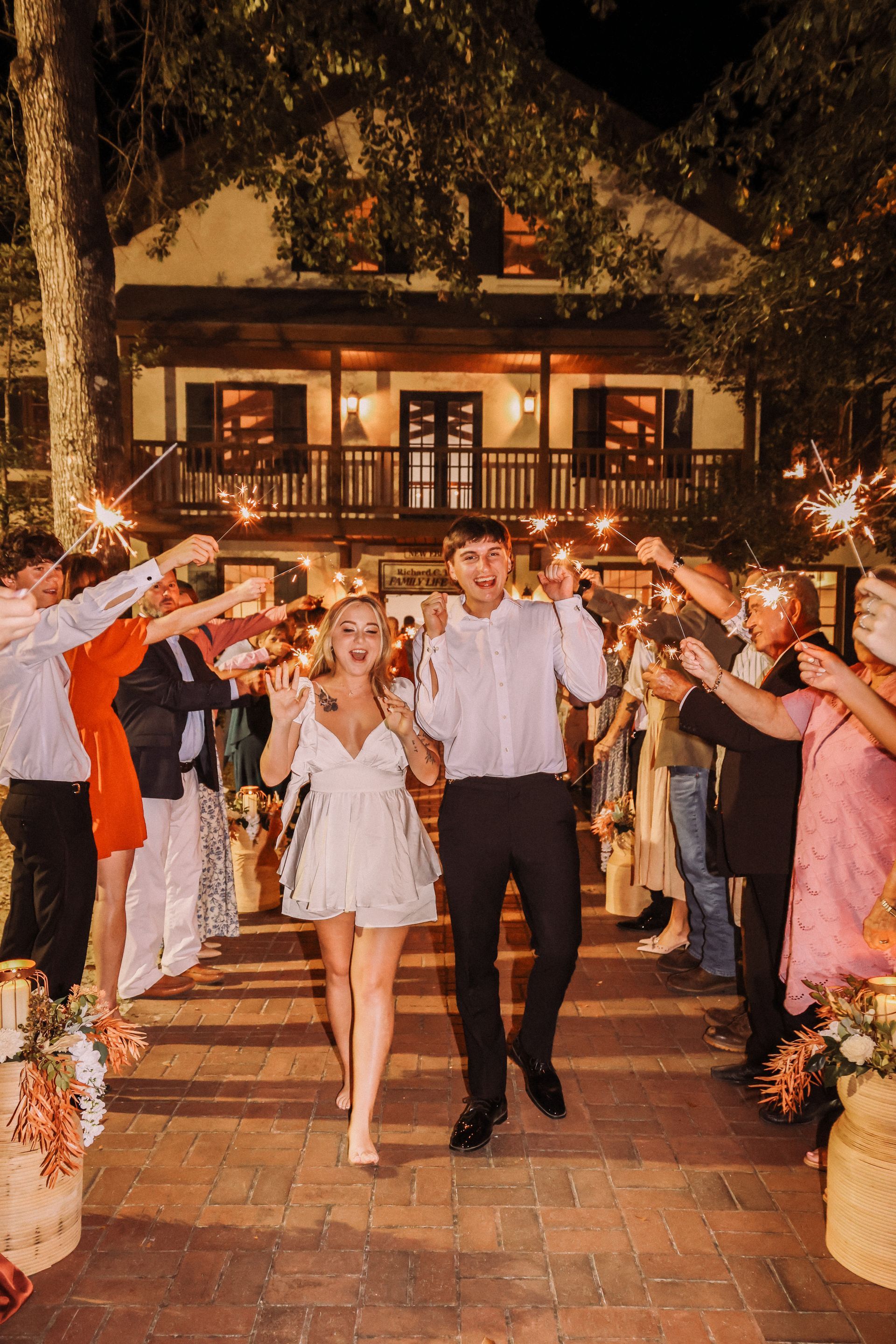 Couple runs through sparkler-lit pathway, celebrating wedding. Guests cheer; rustic building in background.