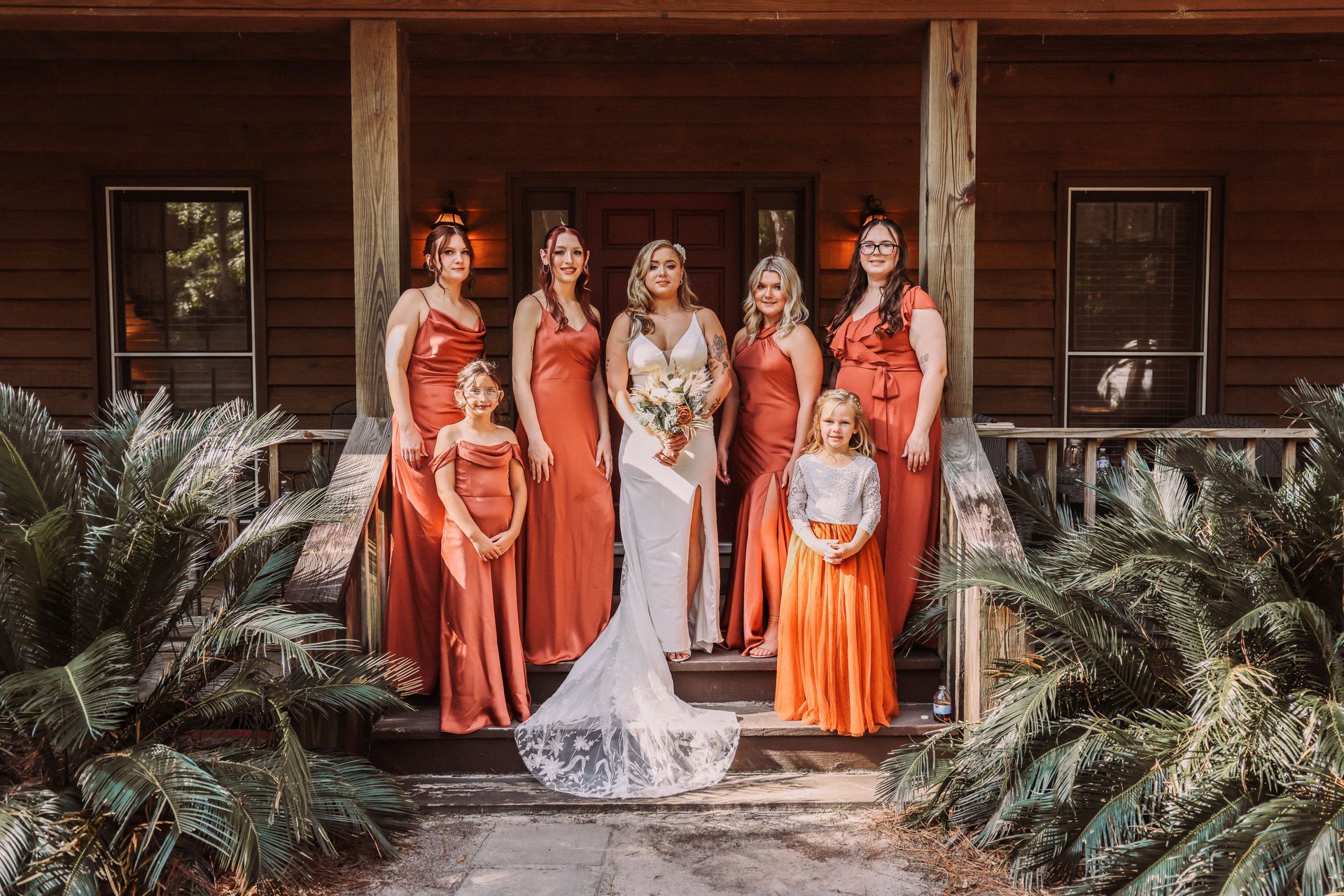 Bride and bridesmaids pose on porch. Bride in white dress, bridesmaids in rust-colored dresses. Outdoor setting.