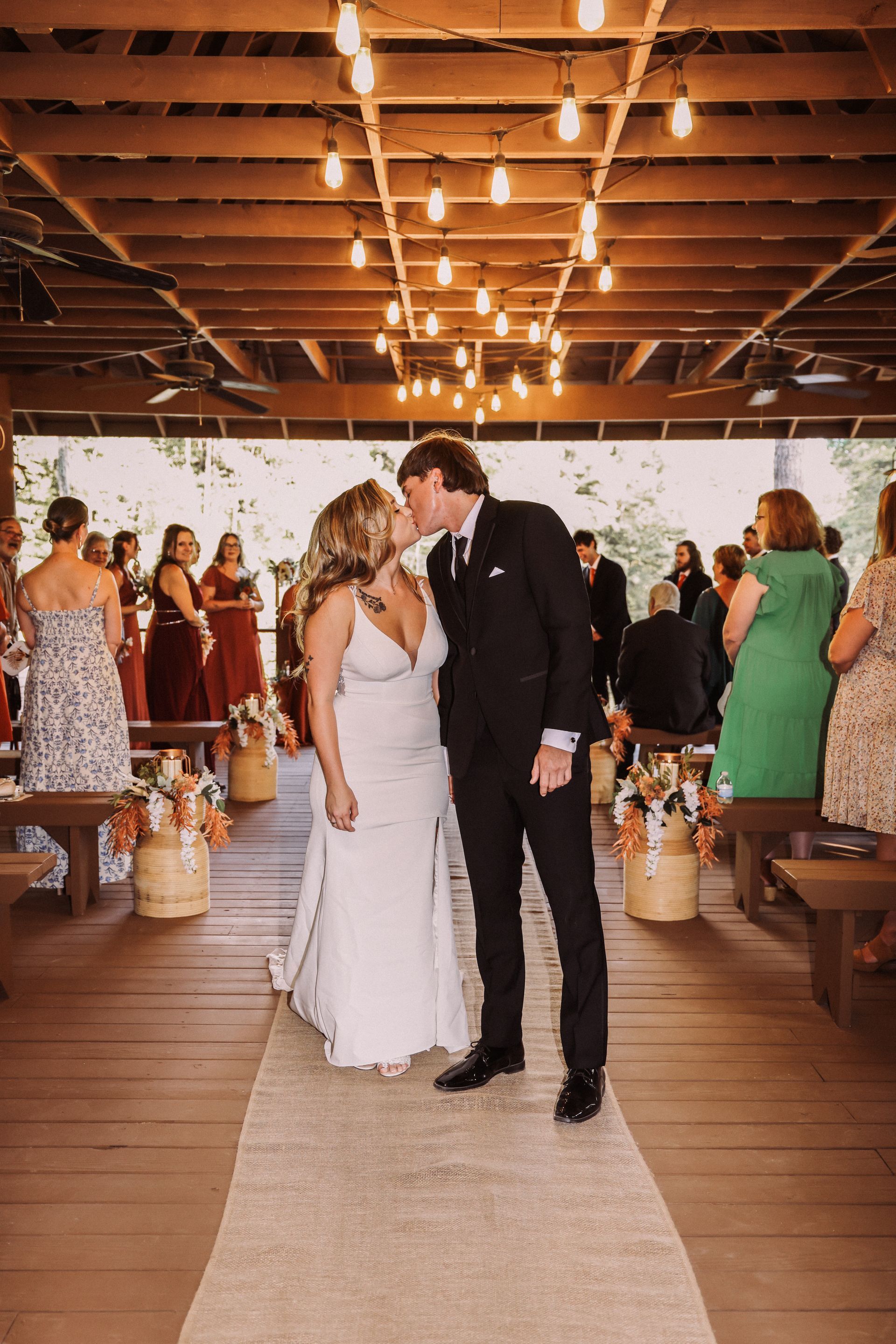 Couple kissing at wedding ceremony, under string lights, on wooden deck.