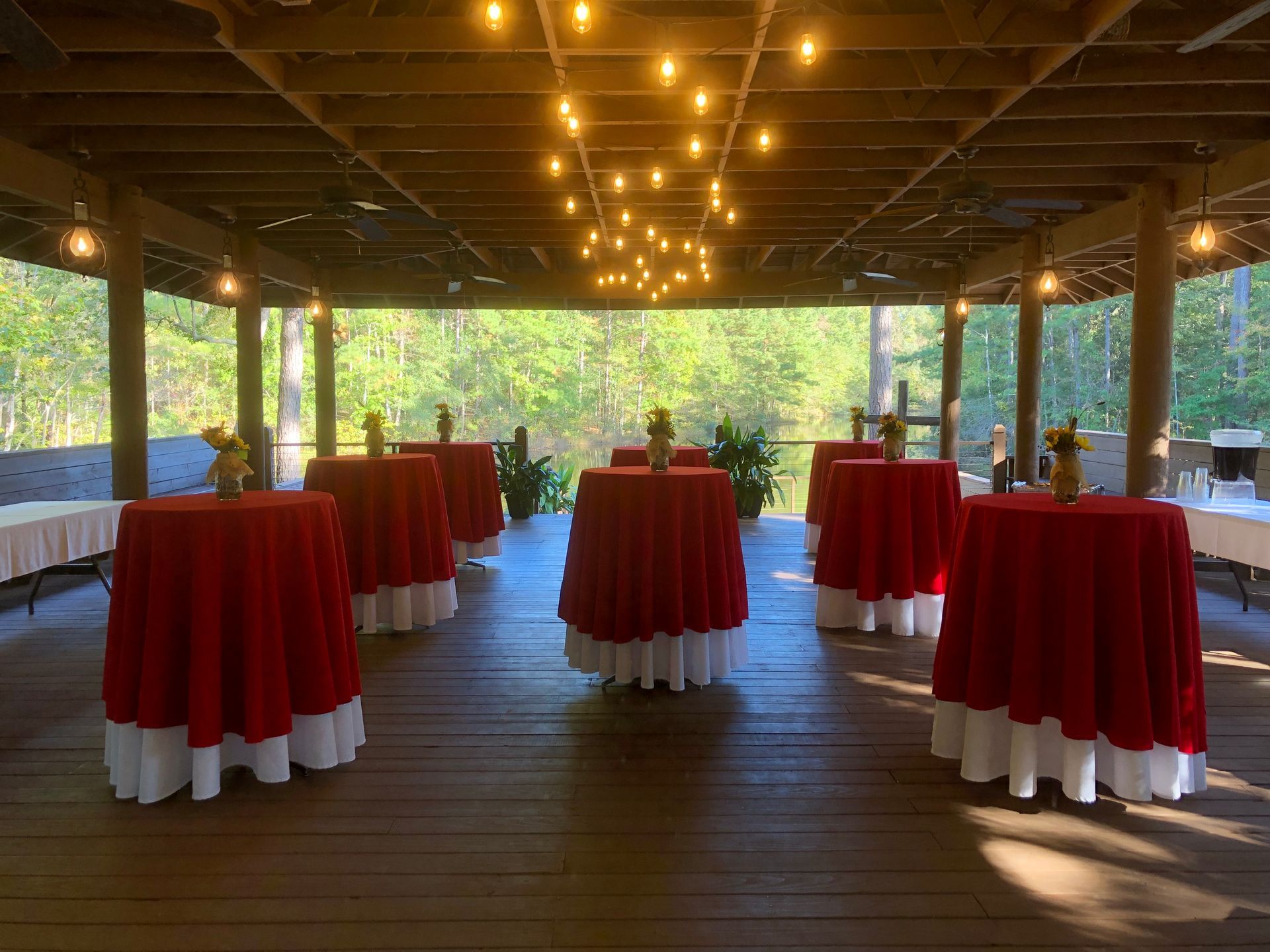 Red and white covered tables set up under a wooden covered patio with string lights, trees in background.