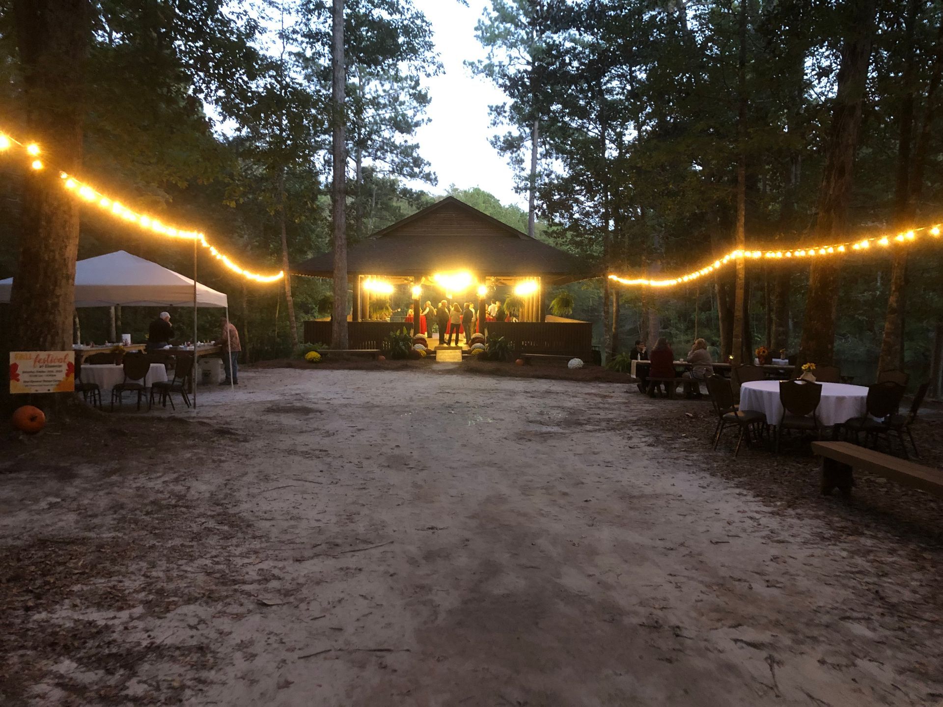 Outdoor event: illuminated pavilion, string lights, tables, and people gathered on a dirt path in a wooded area.