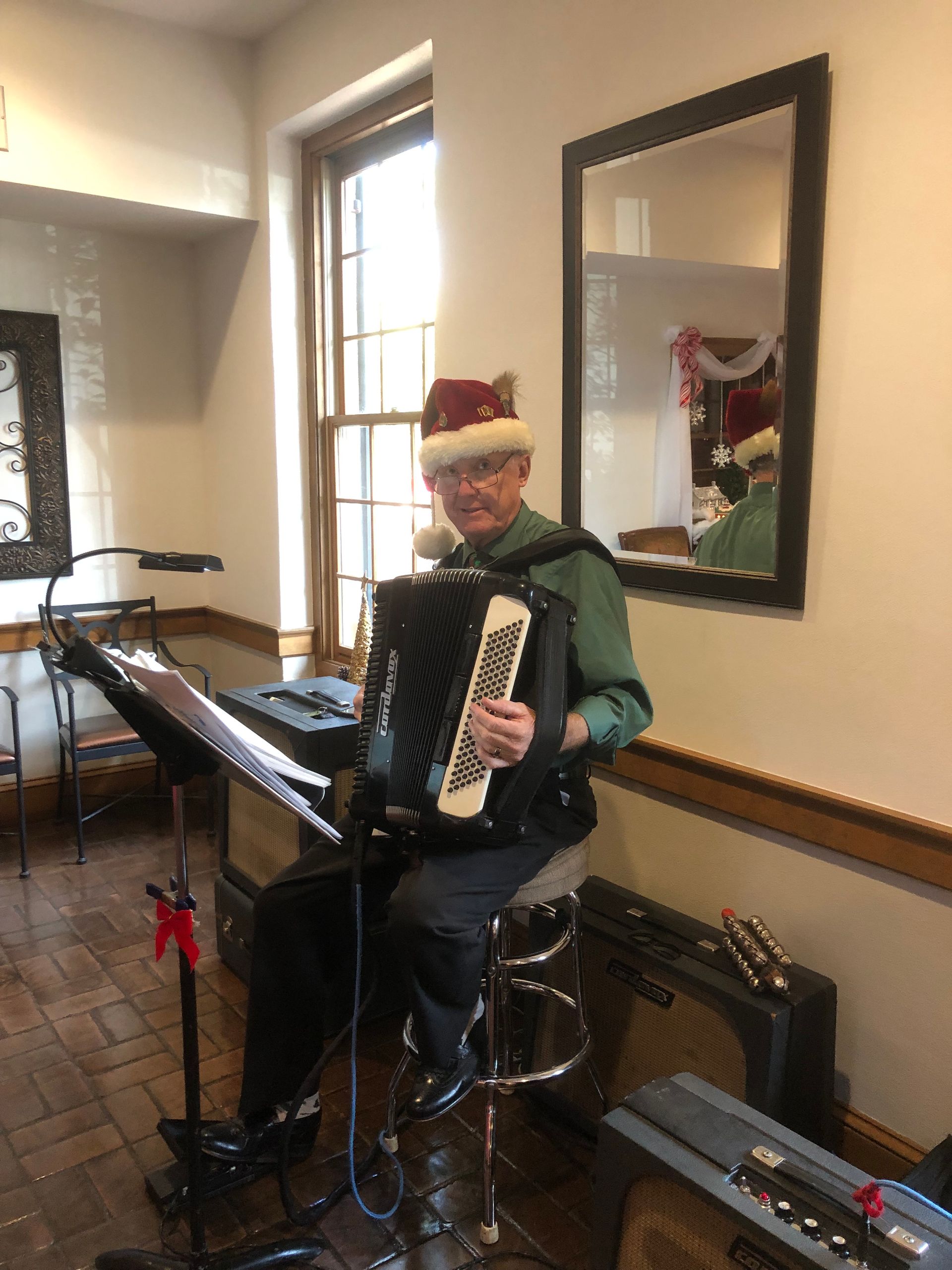 A man in a Santa hat plays an accordion indoors near a window and mirror.