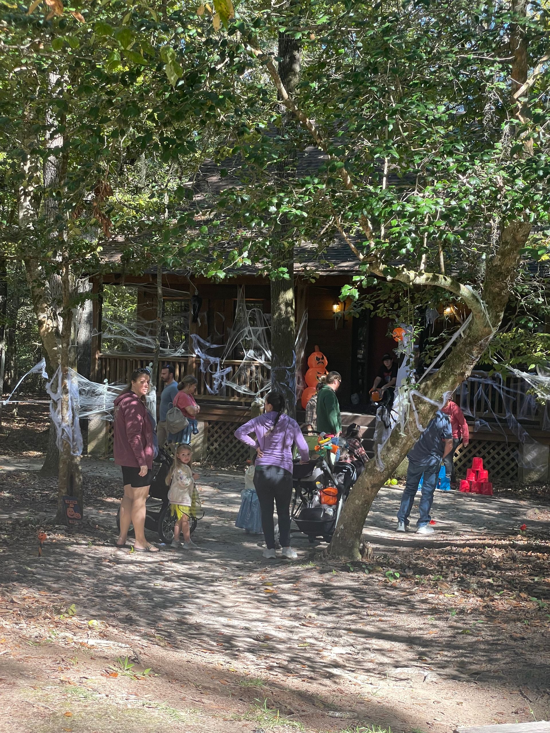 People in costume gather near a decorated house in a wooded area, likely for Halloween.