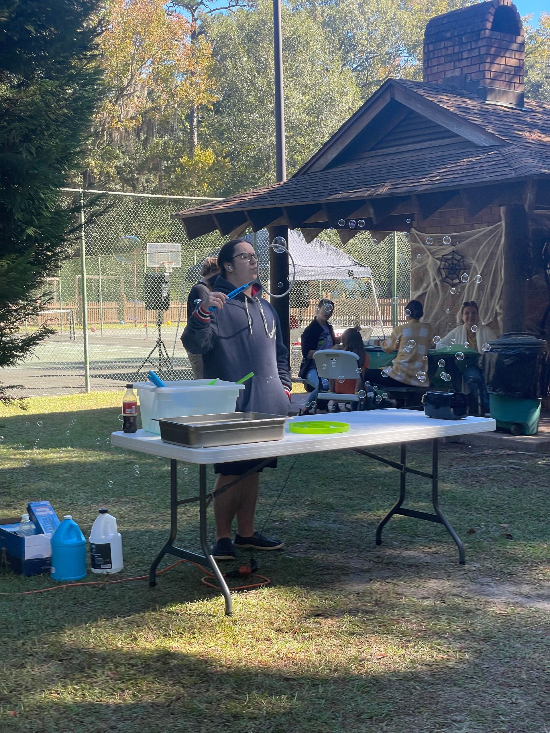 A person stands behind a table with food, an outdoor setting with a building and other people.