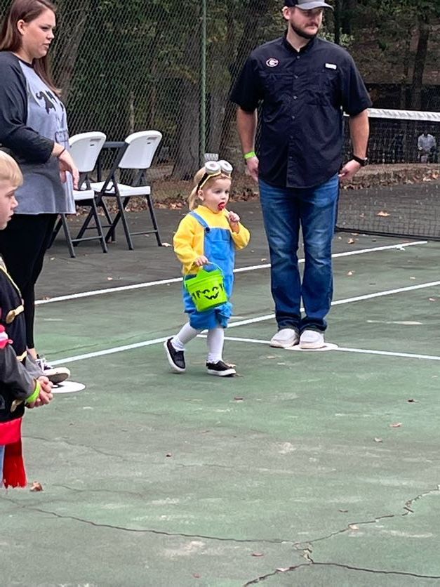 Child in a minion costume, with a candy bucket, on a tennis court, walking towards an adult.