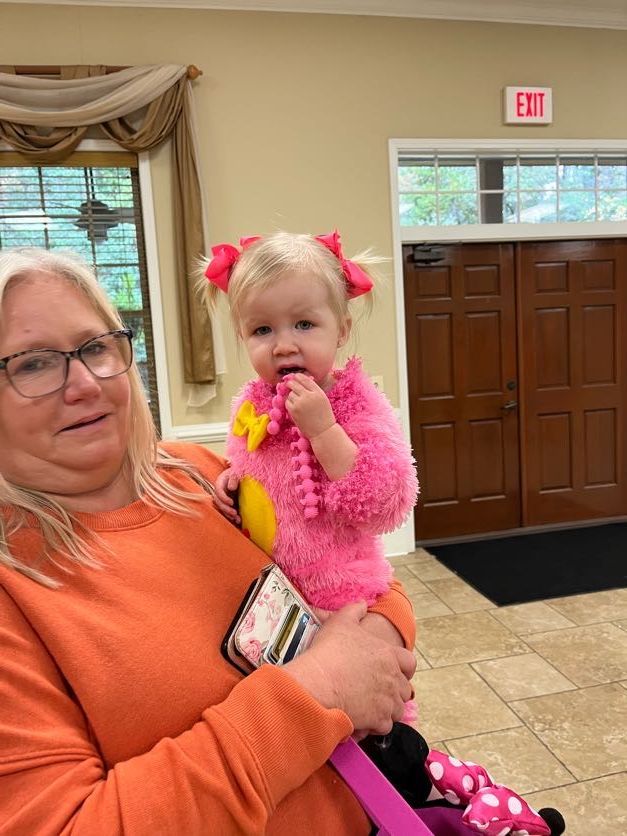 Woman in orange holds toddler in pink costume; interior setting.