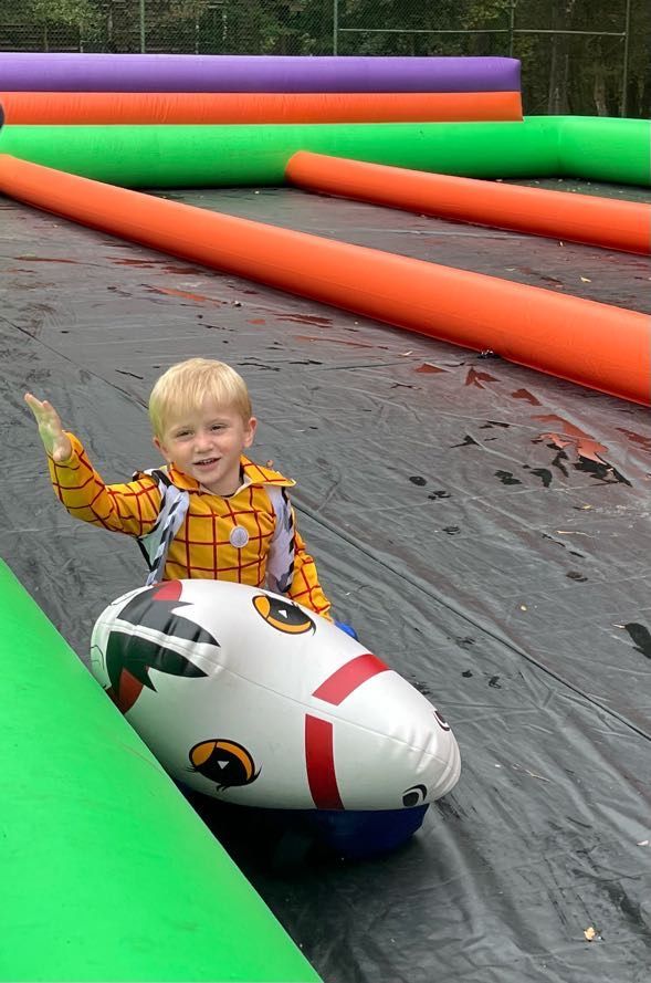 Child in Woody costume waving, riding inflatable toy down a black mat. Green, orange, purple tubes in background.