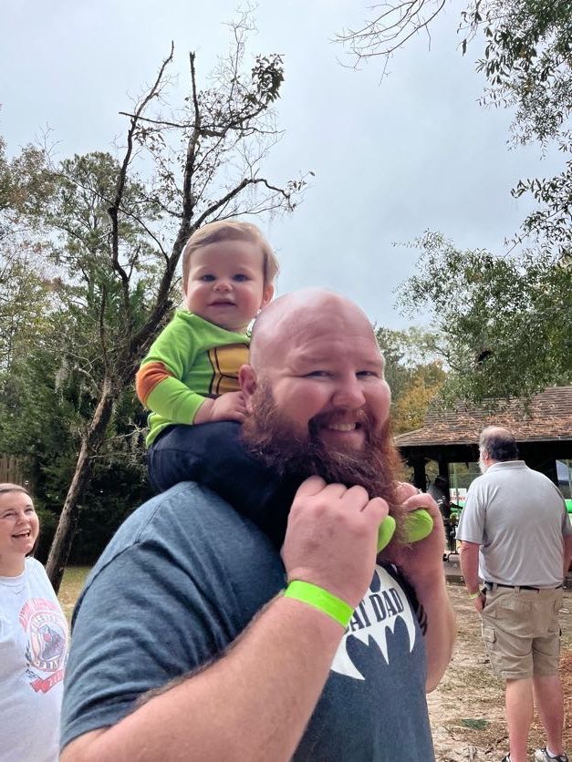 Man with a red beard carries a child on his shoulders. The child smiles, outdoor setting.