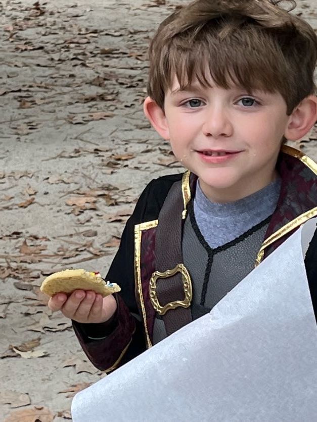 Young boy in costume holding cookie, smiling. Outdoors, wearing a vest and coat.