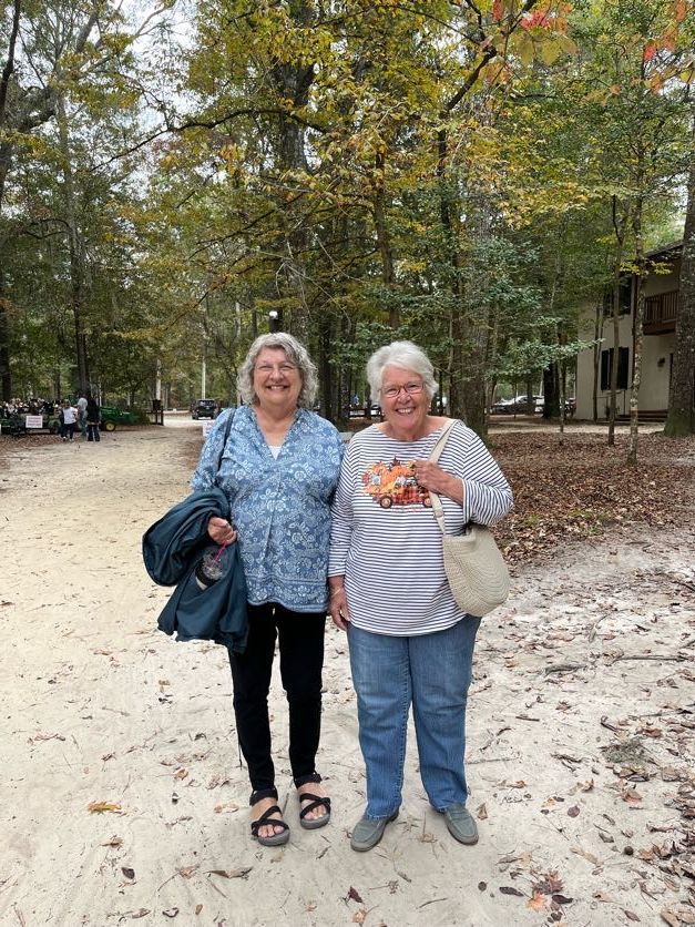 Two women stand on a wooded path. One in blue top, black pants, the other in striped top and jeans.