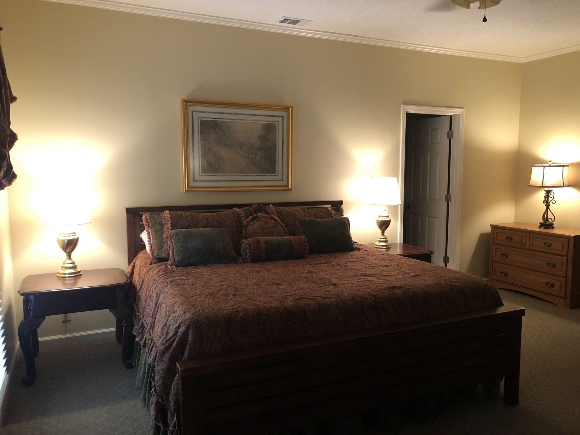 Bedroom with a large bed, nightstands with lamps, a dresser, and a door. Warm lighting and patterned bedding.