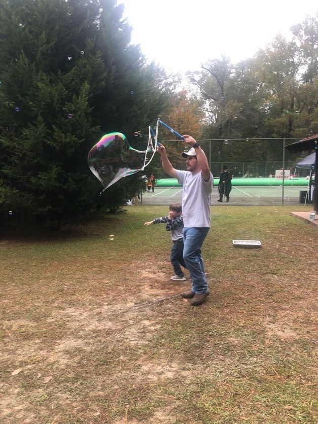 Man and child blowing large bubbles in a grassy yard near a tennis court.