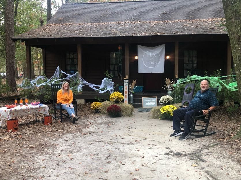 Two people sit outside a decorated cabin with Halloween decorations; fall colors.