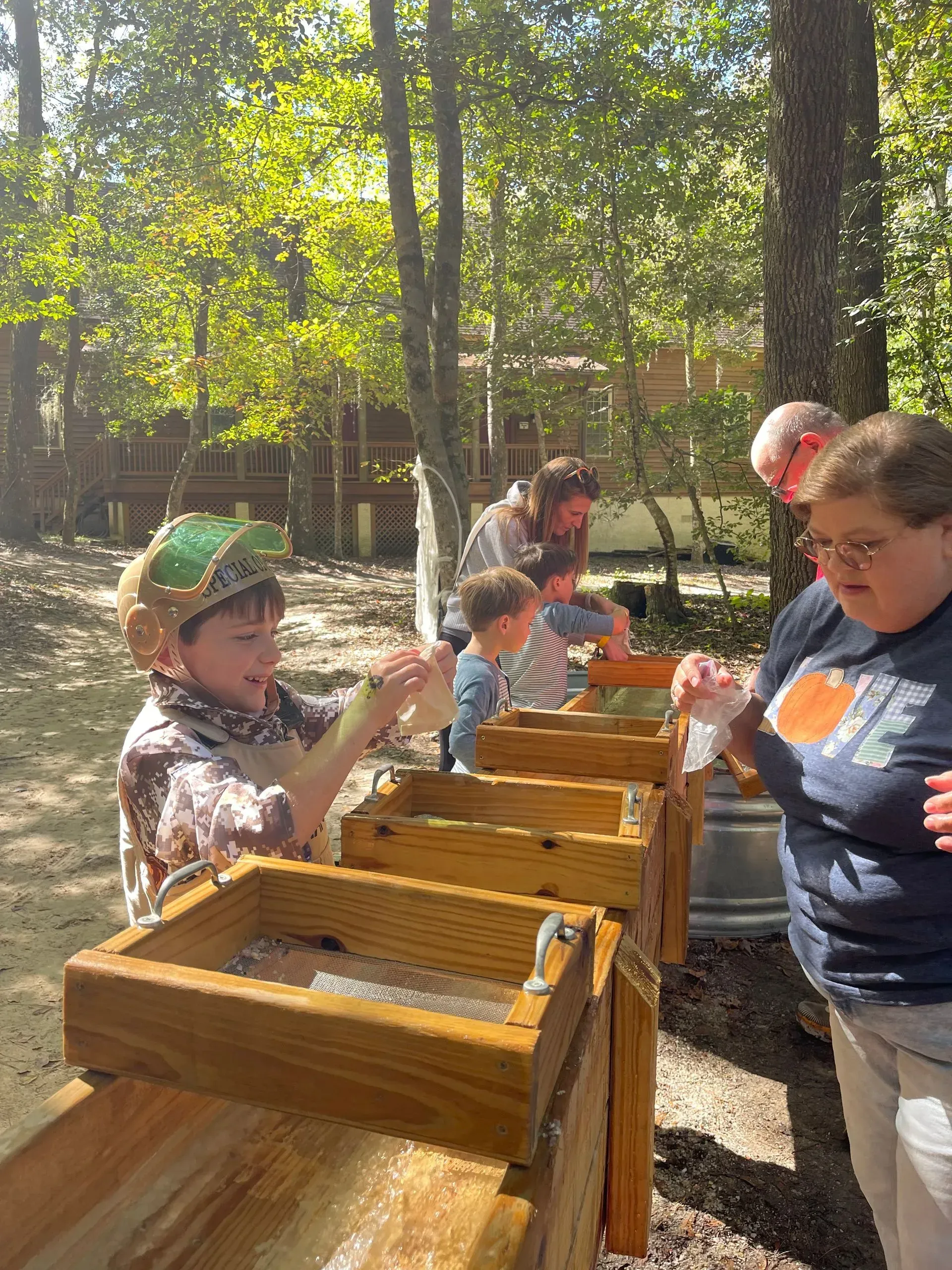 People panning for gems at an outdoor sluice. Smiling child in foreground, other people behind.