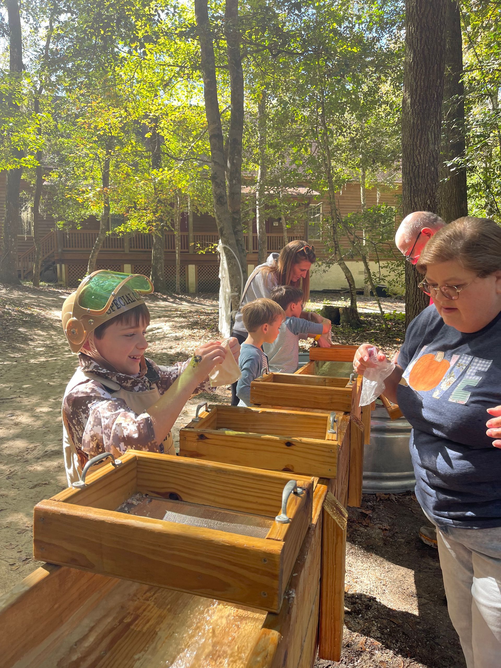 People panning for gems in wooden sluices outdoors. Child smiles, others watch.