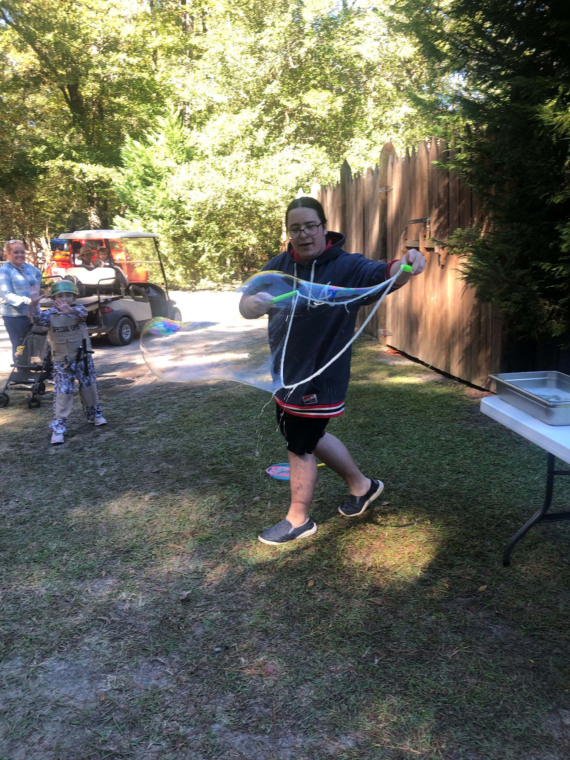 Person blowing bubbles with large wand outdoors; child watches.