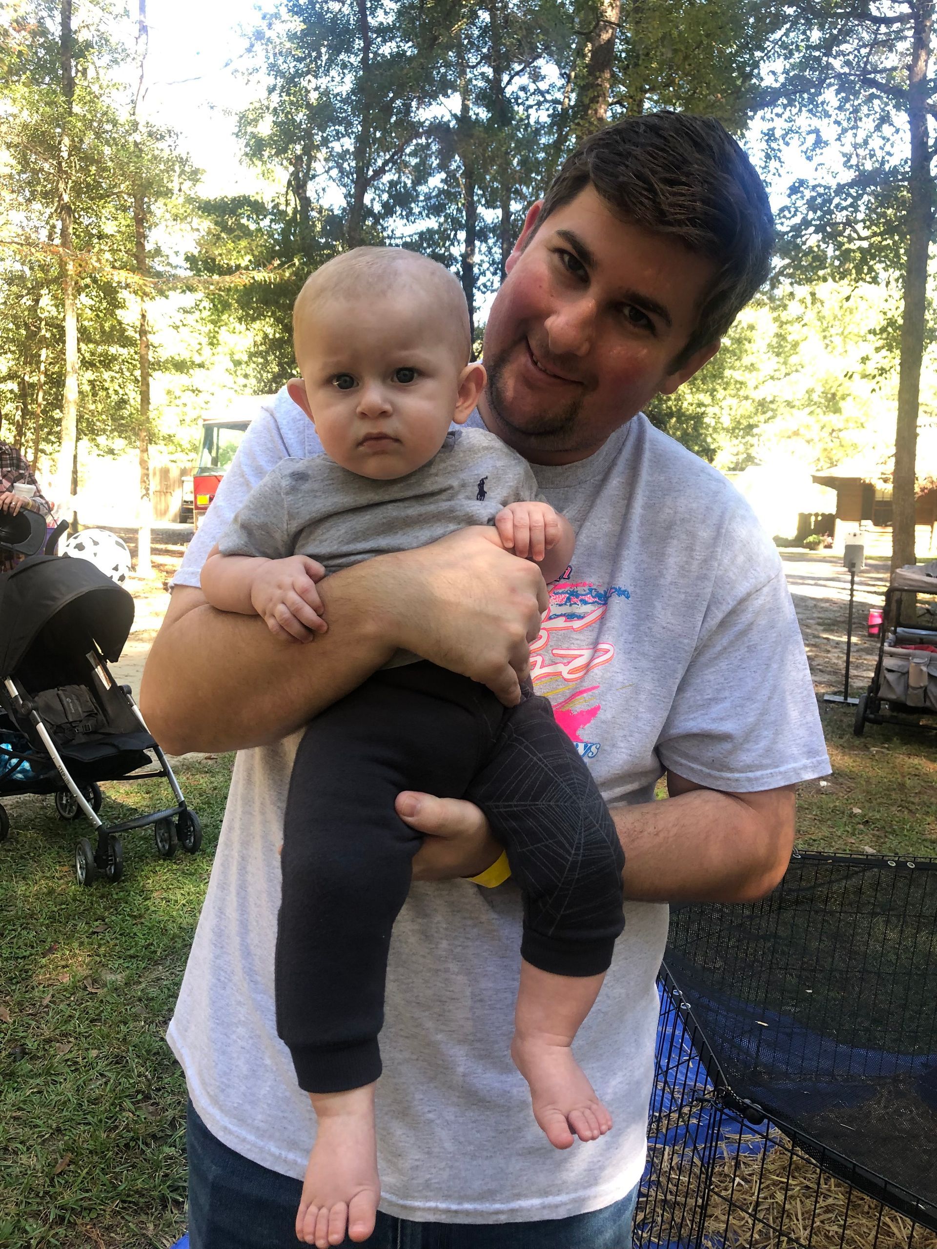Man holding a baby outdoors; both looking at camera. Man wears grey shirt, baby, grey outfit. Green background.