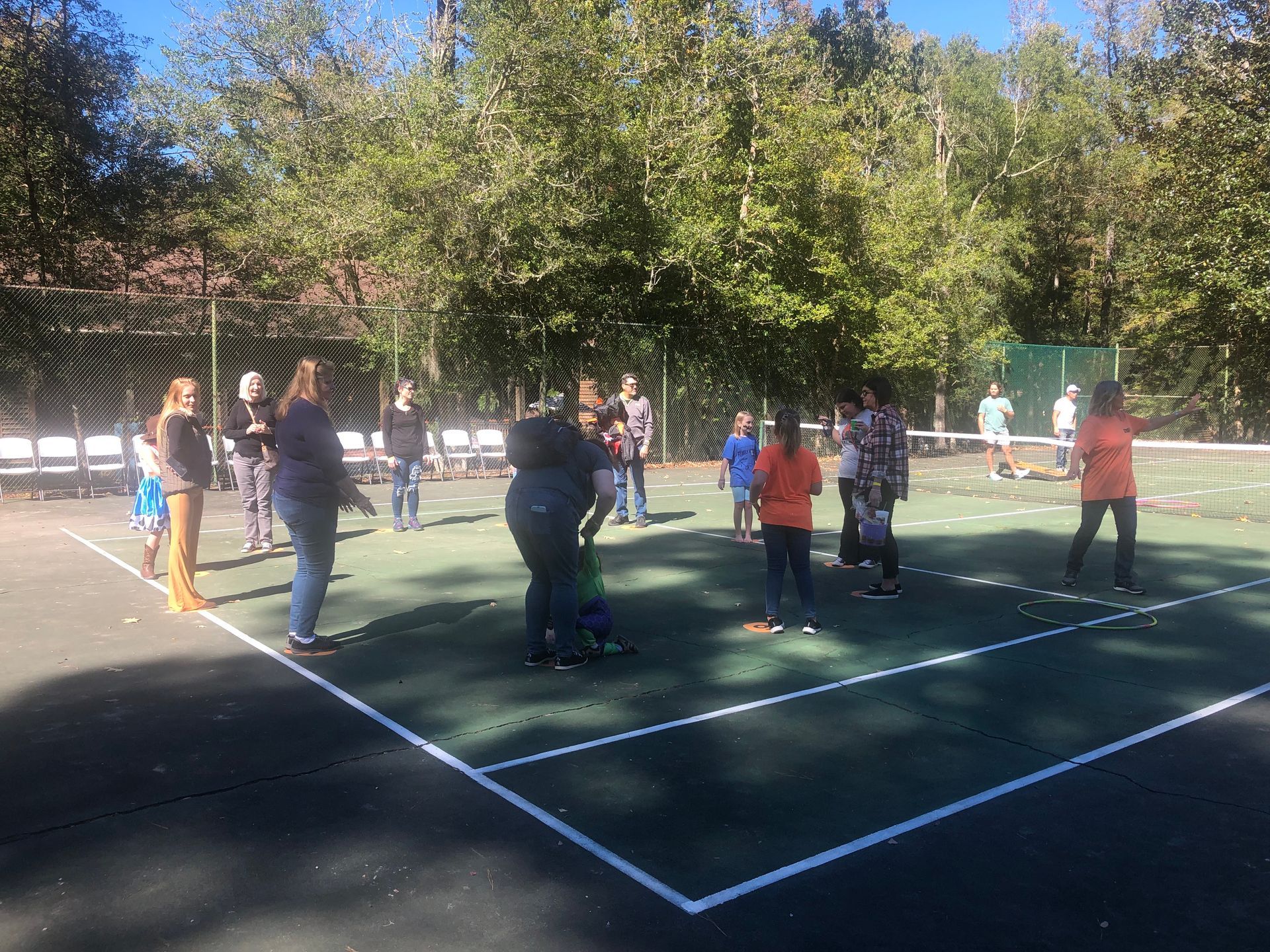 People playing on a green court surrounded by trees, some holding balls, in an outdoor setting.