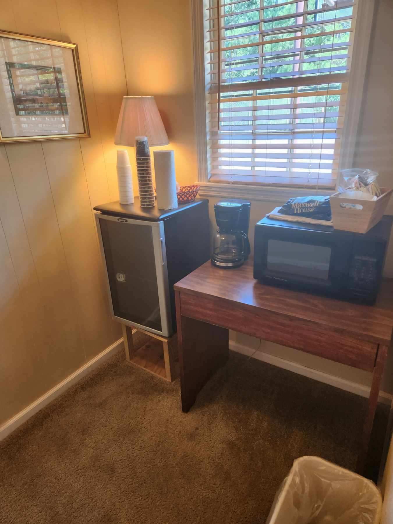 A small room corner with a mini-fridge, lamp, microwave, coffee maker, and a window.