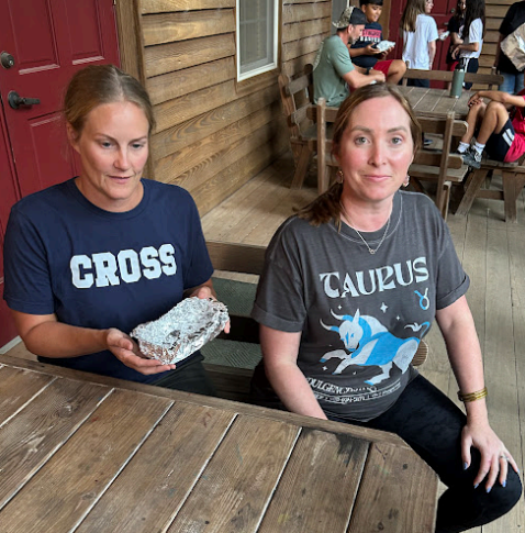 Two women seated at a wooden table on a porch. One holds a rock sample, the other wears a Taurus shirt. Others in background.
