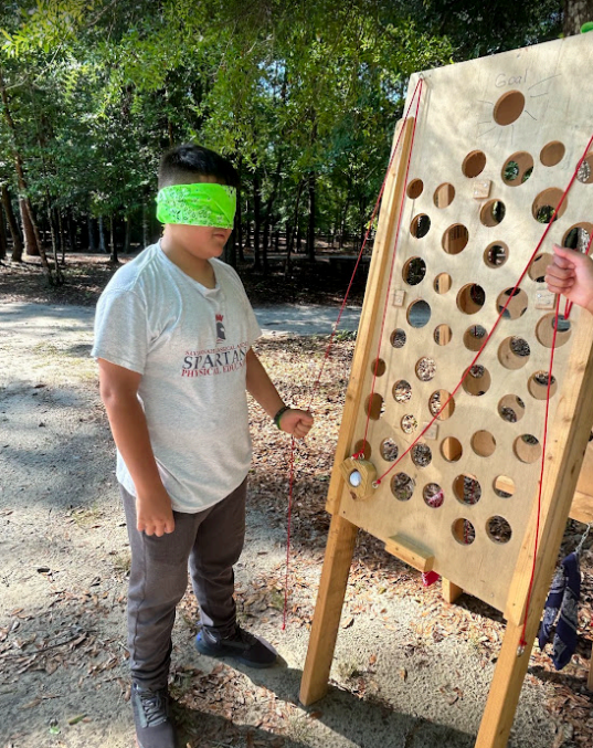Person blindfolded, playing a wooden game with holes and string outdoors.
