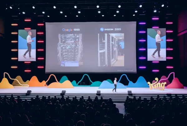 Conference stage with giant central screen, side presenters, and colorful wave graphics above the audience.