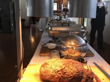 Buffet table with a large baked dish in the foreground and stainless serving trays under warm lights