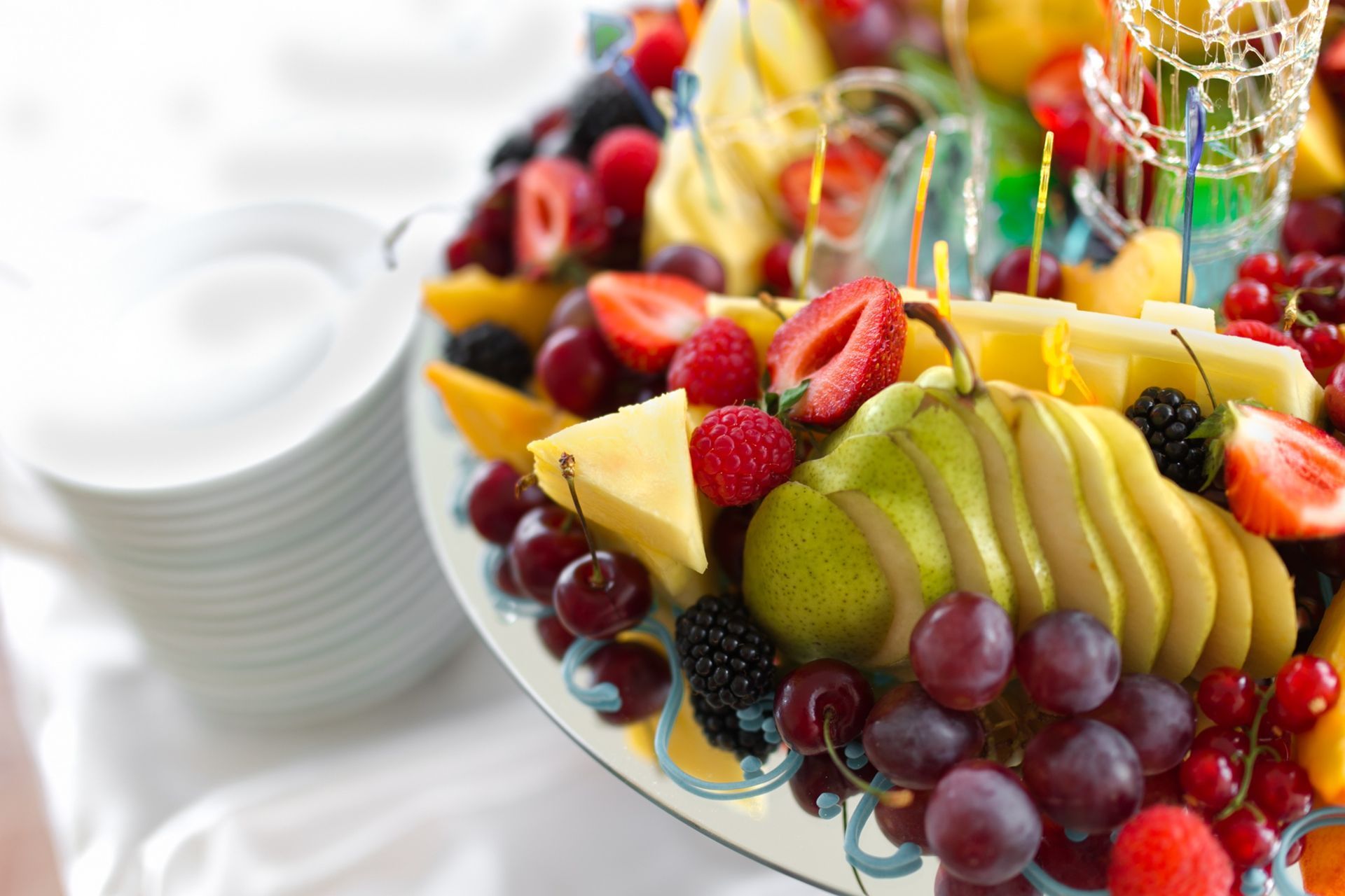 Colorful fruit platter with sliced apples, grapes, strawberries, and pineapple beside stacked white plates