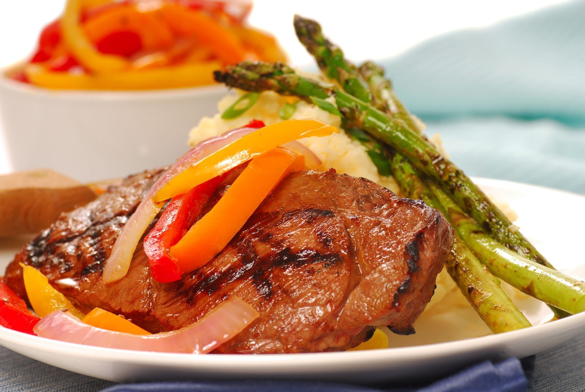 Grilled steak with colorful pepper strips and asparagus on a white plate, with a side dish in the background