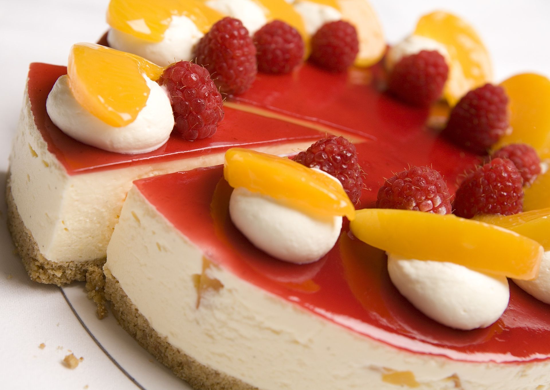 Slice of cheesecake topped with red glaze, raspberries, and peach slices on a white plate