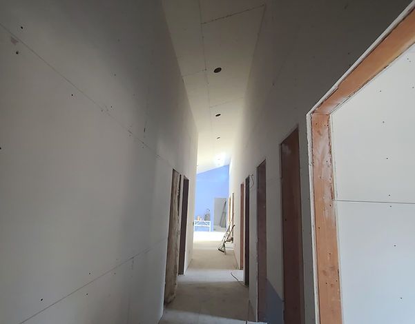 A narrow hallway under construction, with unfinished drywall walls and doorways. Sunlight streams in from the end of the hall.