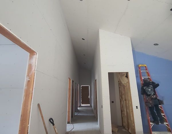 Interior of a building under construction. A worker on a ladder paints a blue wall. Drywall lines a hallway with open doorways.