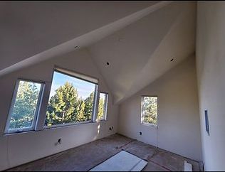 An empty room with sloped ceilings and windows overlooking trees. The room is unfinished with exposed wiring and a neutral color scheme.