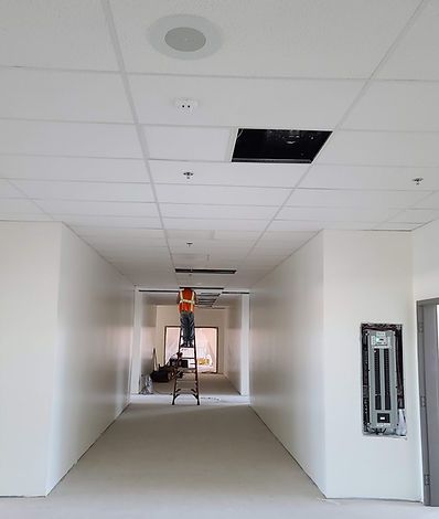 A person on a ladder working in a long, white hallway with a drop ceiling and recessed lighting.