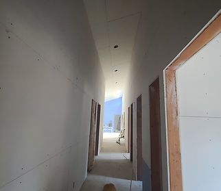 Narrow hallway under construction, walls and ceiling are white drywall. Door frames and trim are brown. Light at the end.