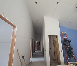 Construction site interior: long hallway with unfinished drywall and worker on a ladder. Blue wall visible.