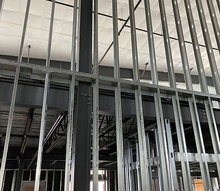 Interior view of a building under construction, showing a steel frame wall and ceiling supports. Grey metal beams are prominent.