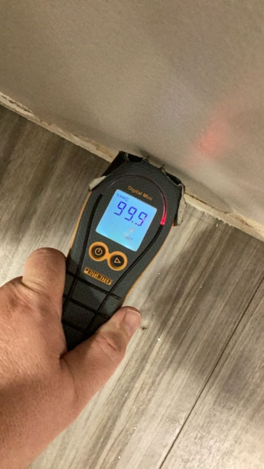 Protimeter moisture meter showing 99.9 reading on saturated drywall near baseboard