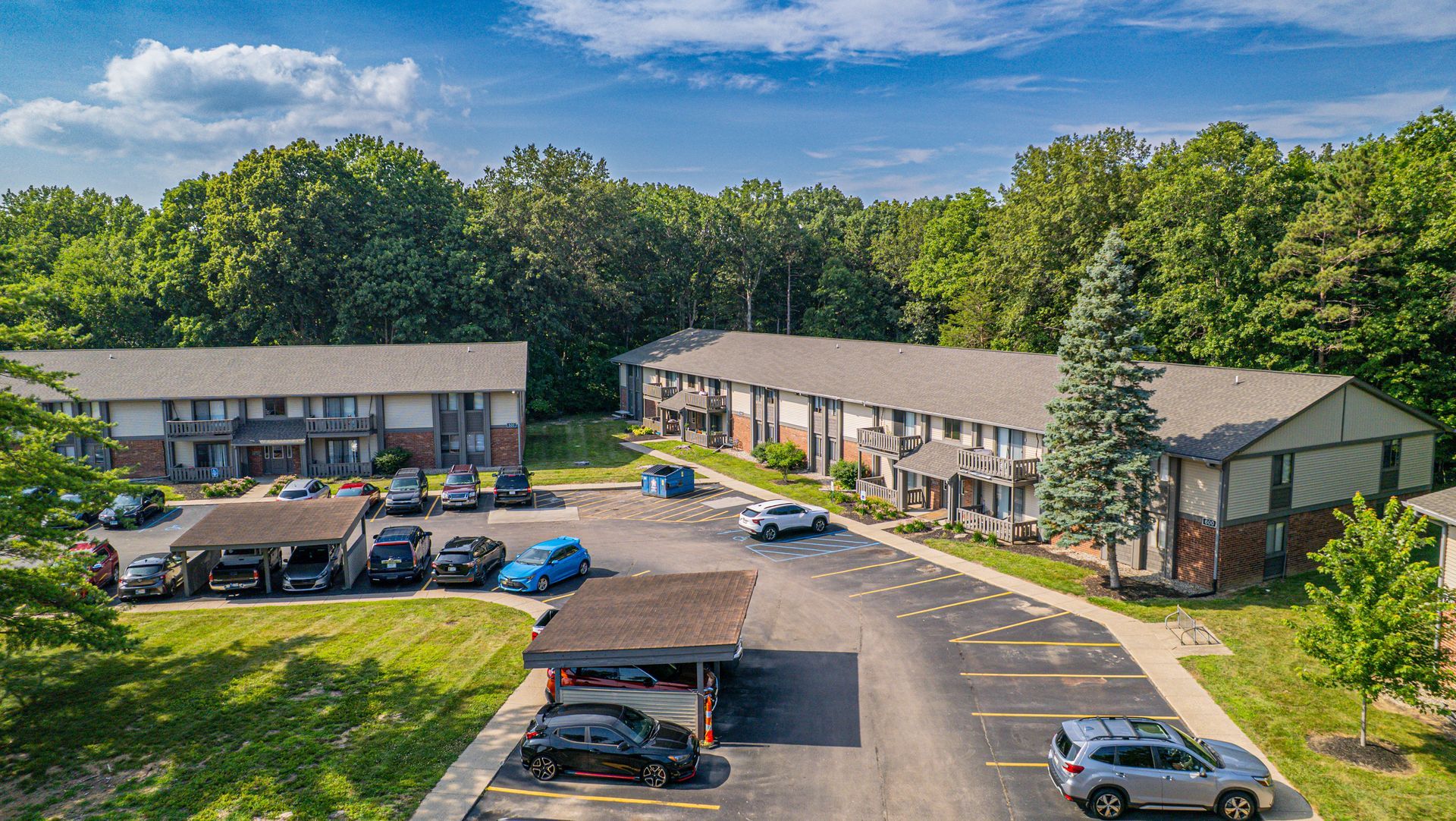 Apartment complex surrounded by trees, with parked cars and covered parking.