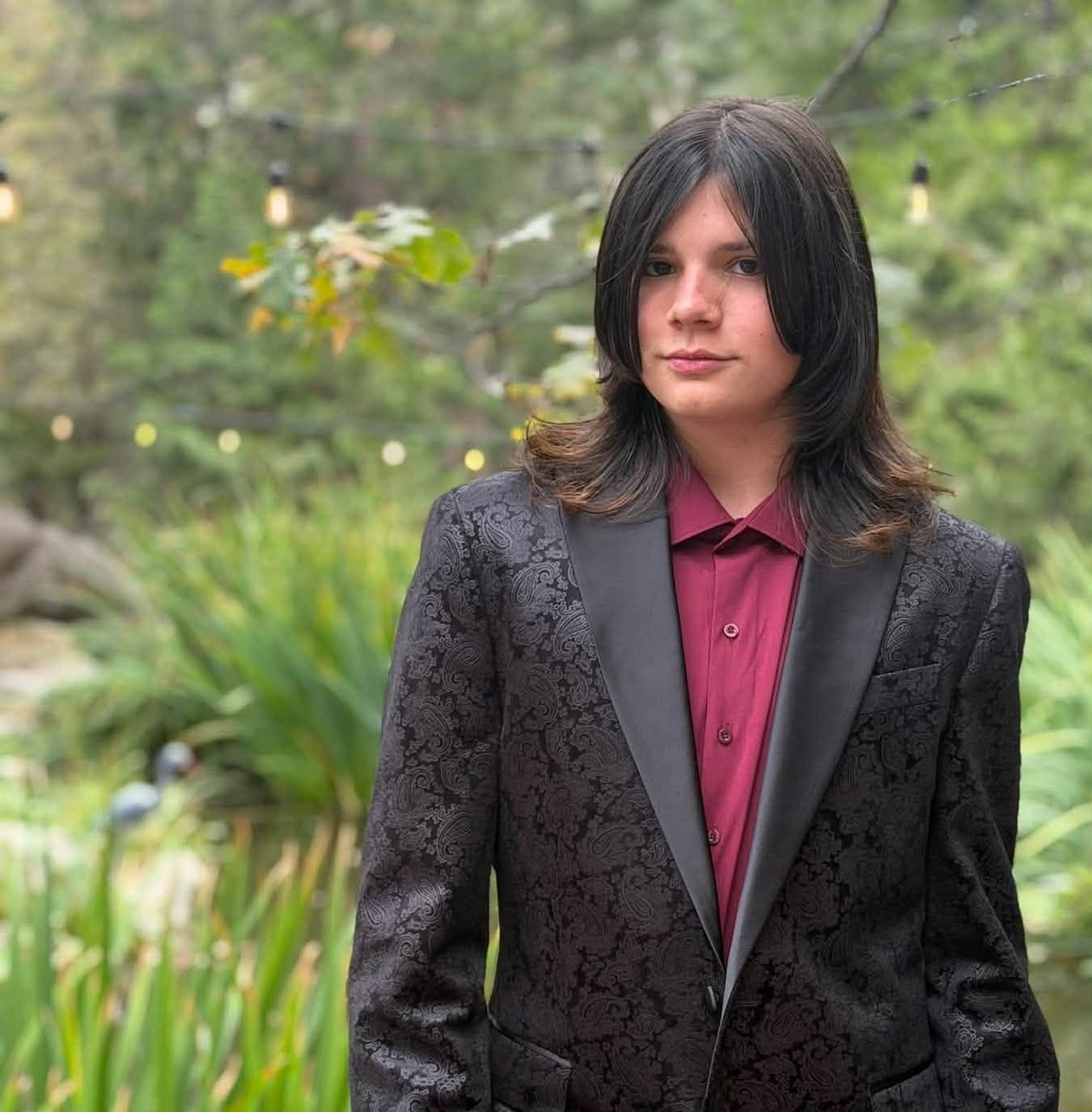 Person in a dark patterned blazer and maroon shirt standing in a garden, looking at the camera.