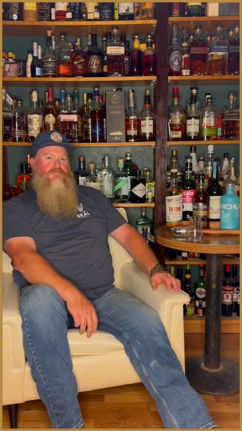 Man sitting in a liquor store aisle beside shelves of bottles and a small table, wearing a cap and jeans.