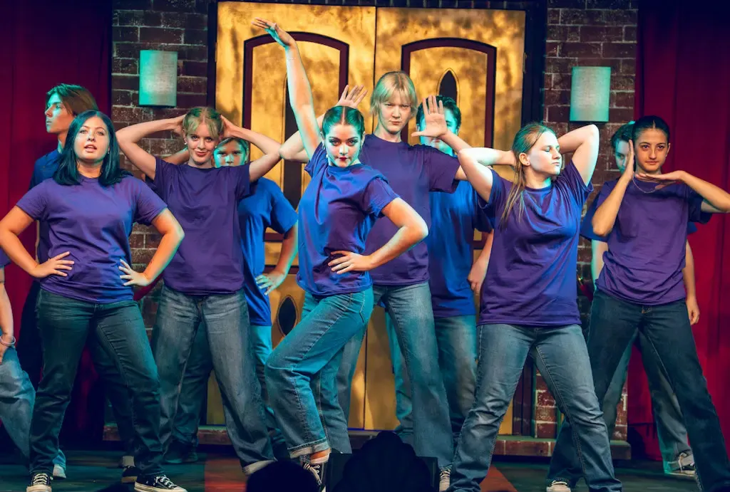 Group of dancers in purple shirts and jeans posing on stage with gold backdrop and colorful lighting
