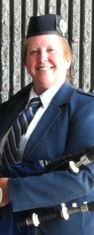 Woman in a blue uniform smiles, holding bagpipes. She wears a hat and tie. Gray background.