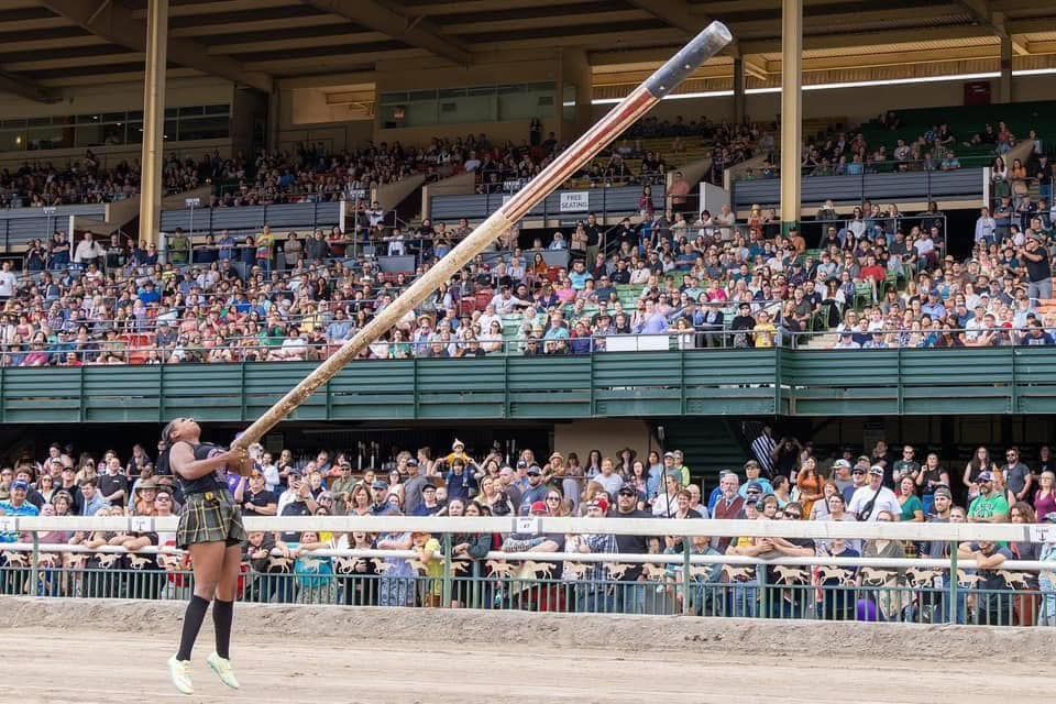 Woman playing a large alphorn in an outdoor arena with a crowd of spectators.