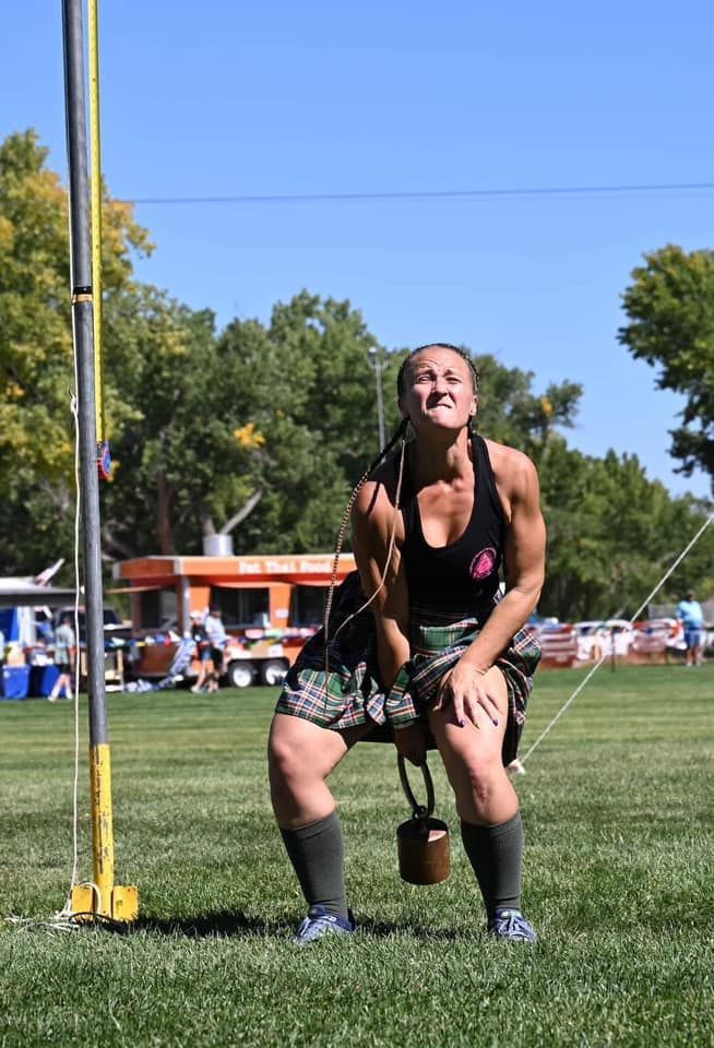Woman in a kilt lifting a weight overhead at a Highland games event.