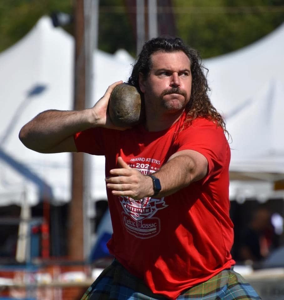 Man in red shirt throws stone, wearing a kilt, in a competition setting.