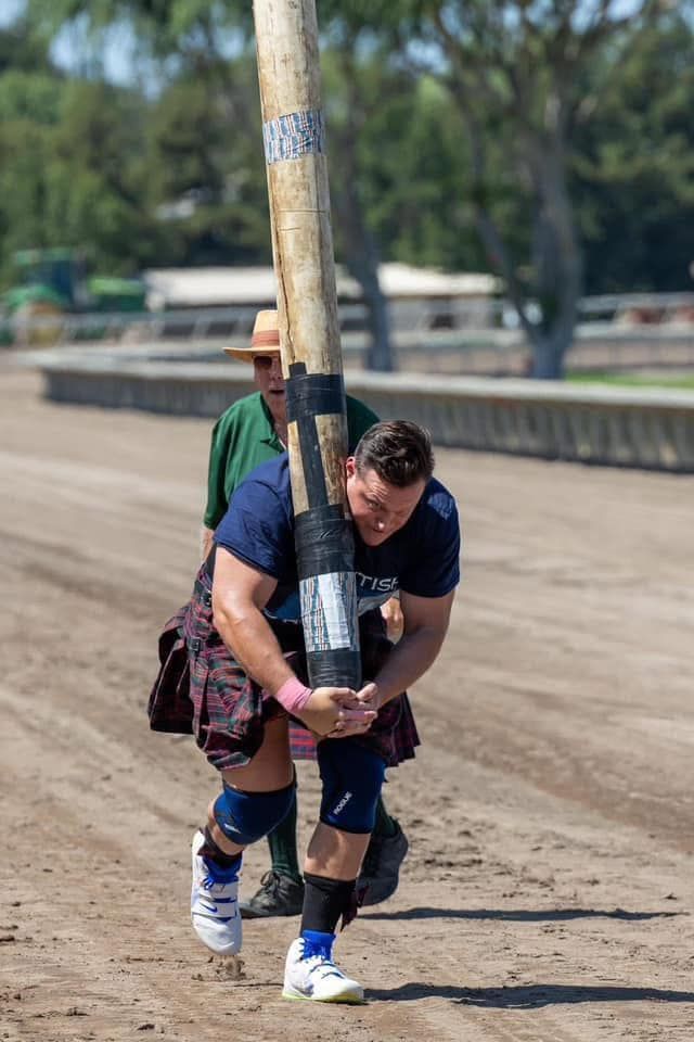 Man in kilt carries a heavy log over his shoulder during a Scottish Highland Games event on dirt.