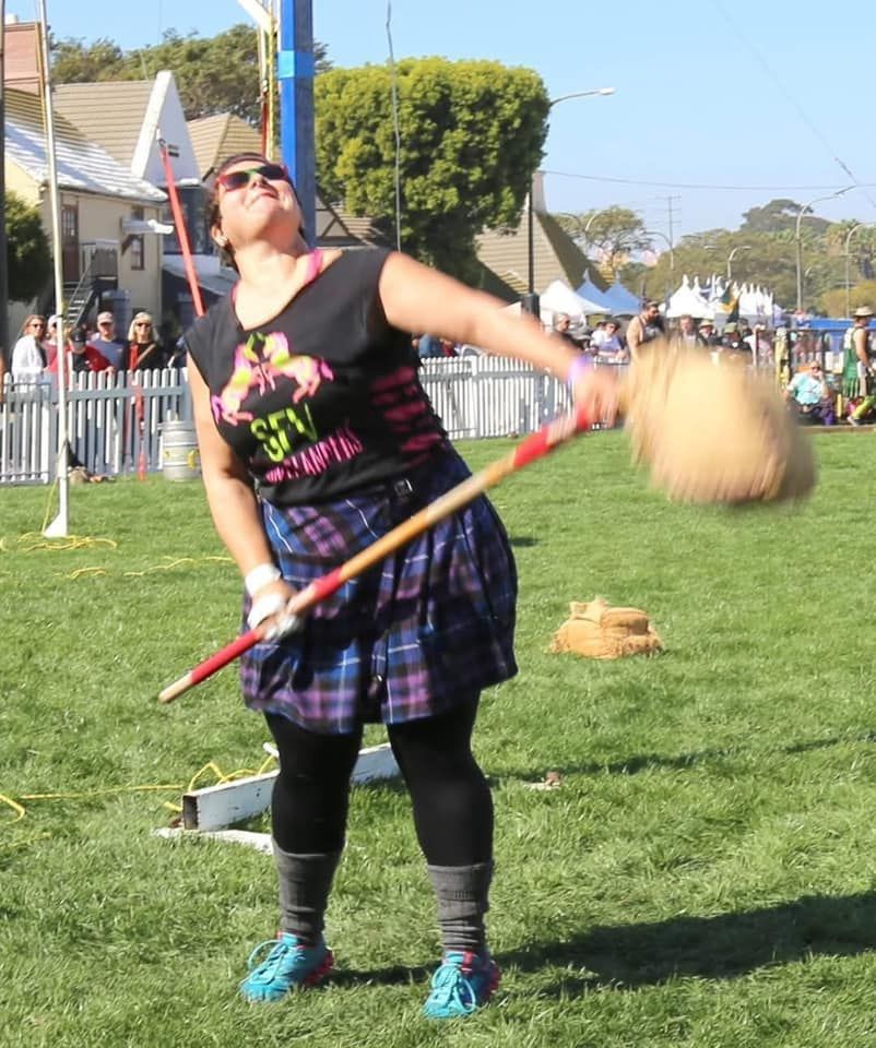 Woman throwing a caber at an outdoor Scottish festival. She wears a black shirt, plaid skirt, and is outdoors on grass.